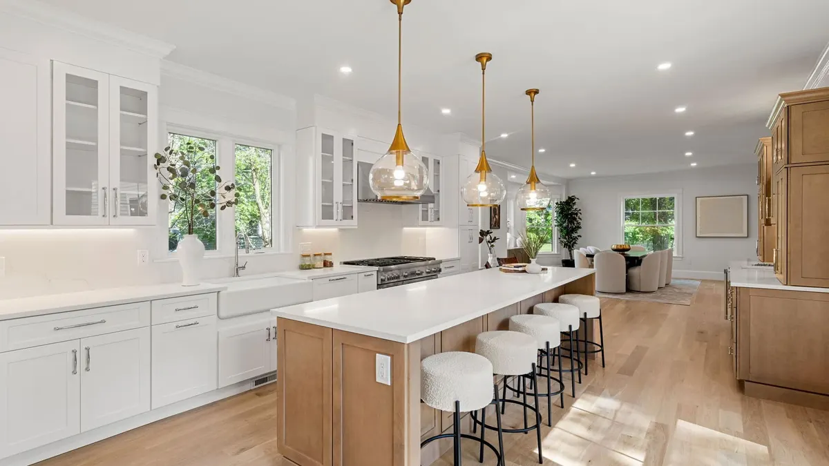 A warm white-and-wood kitchen with a natural-oak island, brass pendants, and glass-front cabinetry open to the living room, a Bay State Kitchen Gallery project in Newton